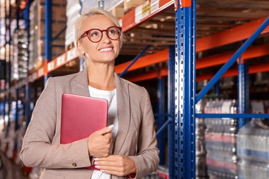 Happy manager holding modern tablet in warehouse with lots of products. Space for text
