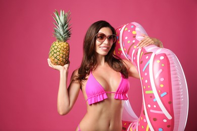 Beautiful woman in stylish bikini with inflatable ring and pineapple on crimson background