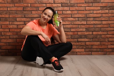 Happy overweight woman with bottle of water sitting near brick wall indoors, space for text