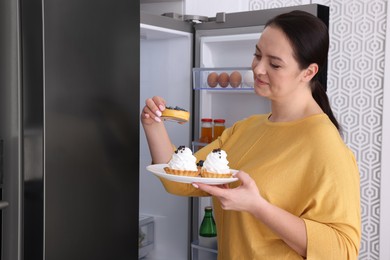 Happy overweight woman with cakes near fridge in kitchen