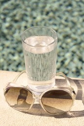 Photo of Stylish sunglasses and glass of water near outdoor swimming pool on sunny day