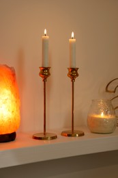 Pair of beautiful candlesticks, scented candle and salt lamp on white shelf