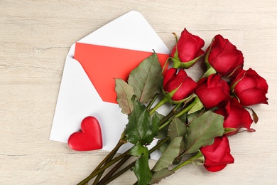 Beautiful red roses, envelope with love letter and decorative heart on white wooden background, flat lay. Valentine's Day celebration