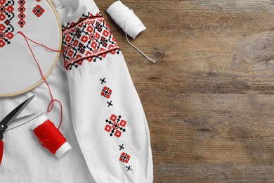 Photo of Shirt with red embroidery design in hoop, scissors and threads on wooden table, flat lay. National Ukrainian clothes