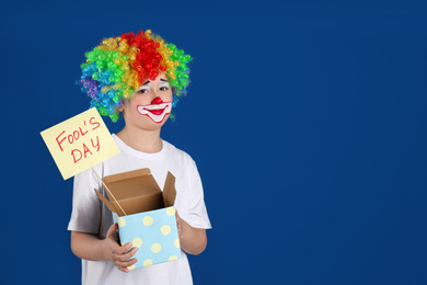 Preteen boy with clown makeup and wig holding sign APRIL FOOL'S DAY on blue background, space for text