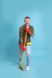 Handsome young man with floor brush singing on light blue background