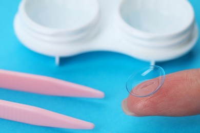 Woman holding contact lens near case and tweezers on light blue background, closeup