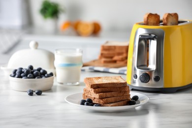 Yellow toaster with roasted bread slices, blueberries and glass of milk on white marble table