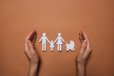 Woman protecting paper family figures on brown background, top view. Insurance concept