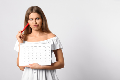 Pensive young woman holding calendar with marked menstrual cycle days on light background. Space for text