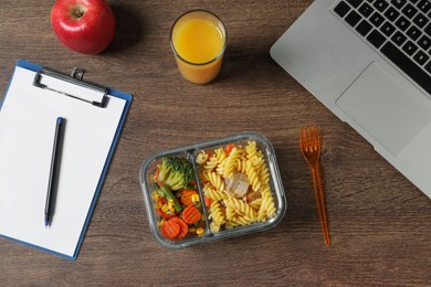 Photo of Container of tasty food, fork, laptop, glass of juice, apple and clipboard on wooden table, flat lay. Business lunch