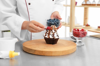 Female pastry chef decorating cupcake with berries at table in kitchen, closeup