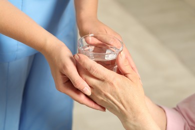 Caretaker giving glass of water to elderly woman indoors, closeup