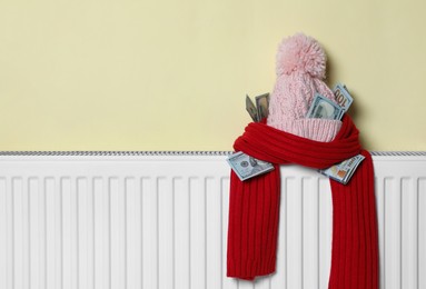 Knitted hat and scarf with money on heating radiator near beige wall, space for text. Energy crisis concept