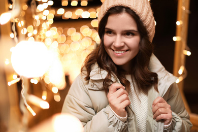 Beautiful young woman near festive lights outdoors. Winter vacation