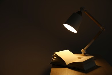Photo of Stylish modern desk lamp and open book on wooden table in darkness, space for text