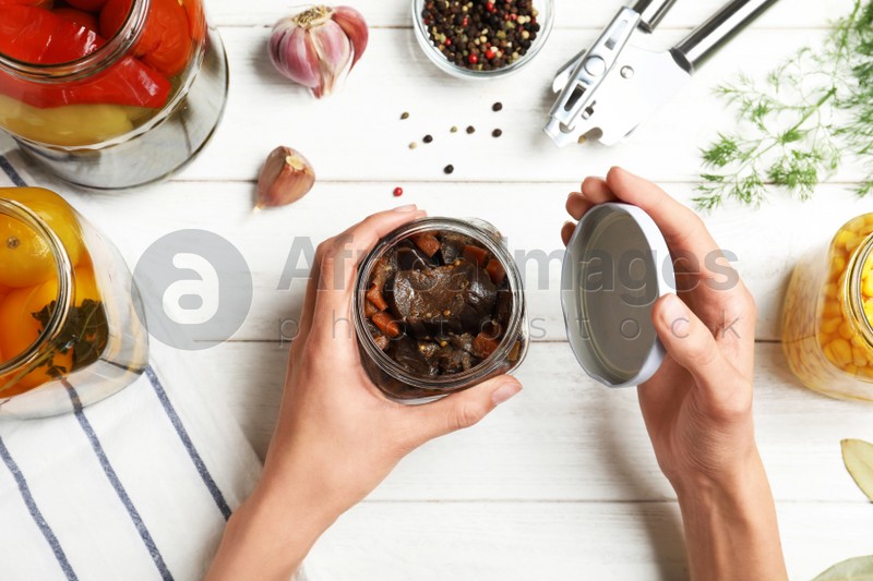 Woman opening jar with pickled eggplants at white wooden table, top view