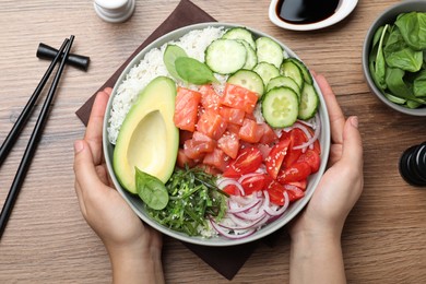 Woman holding delicious poke bowl with salmon and vegetables at wooden table, top view