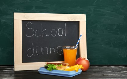 Lunch box with food and small blackboard on wooden table near green chalkboard