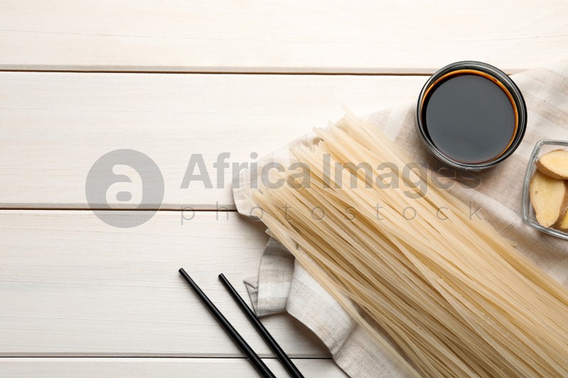 Photo of Uncooked rice noodles, soy sauce, cut ginger and chopsticks on white wooden table, flat lay. Space for text