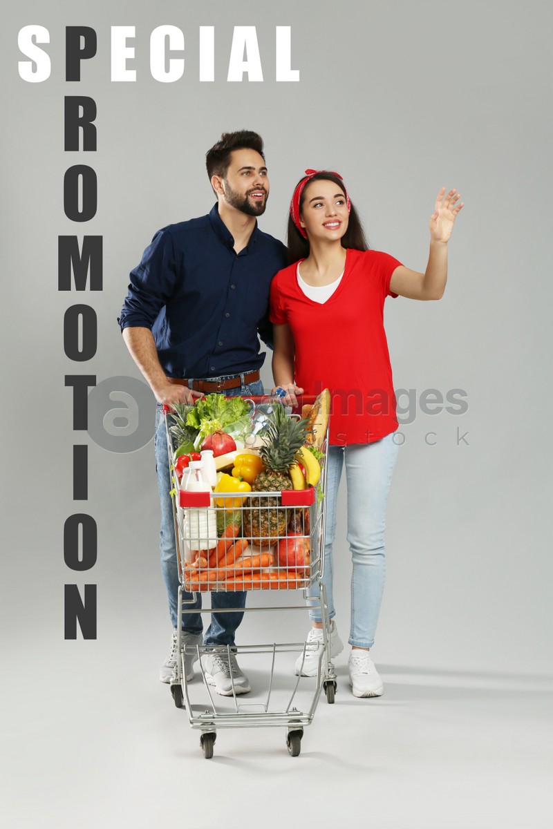 Image of Special promotion. Young couple with shopping cart full of groceries on grey background