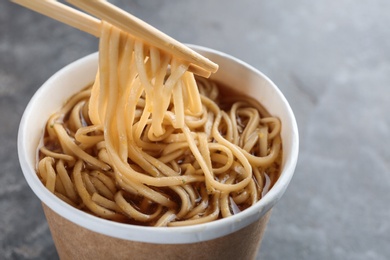Paper cup of instant noodles with sauce and chopsticks on grey background, closeup. Space for text