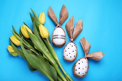 Photo of Easter bunnies made of craft paper and eggs near beautiful tulips on light blue background, flat lay