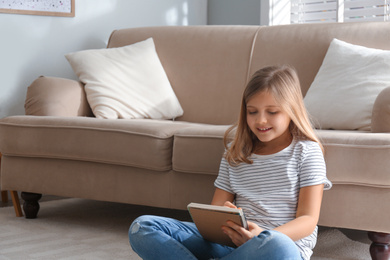 Cute little girl with notebook near couch at home. Space for text