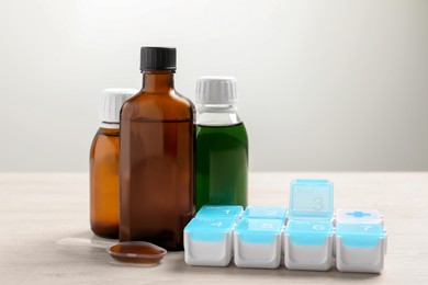 Photo of Bottles of syrup, dosing spoon and weekly pill organizer on white table against light grey background. Cold medicine