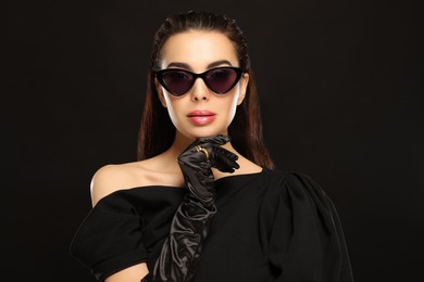 Photo of Portrait of beautiful young woman in sunglasses and elegant evening gloves on black background
