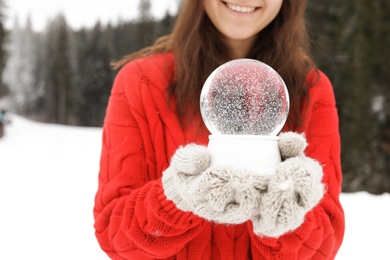 Photo of Woman with knitted mittens holding snow globe outdoors, closeup