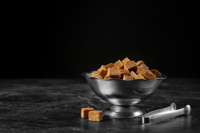 Metal bowl with brown sugar cubes and tongs on black table. Space for text