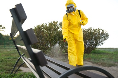 Person in hazmat suit disinfecting bench in park with sprayer. Surface treatment during coronavirus pandemic