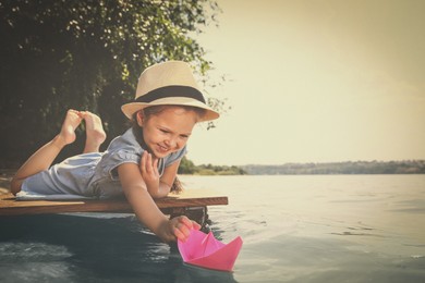 Cute little girl playing with paper boat on wooden pier near river. Retro photo effect 