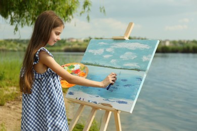 Little girl painting scenery on easel near lake