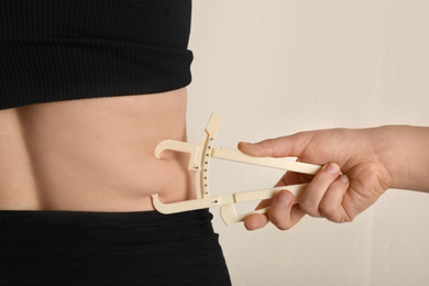 Nutritionist measuring woman's body fat layer with caliper on beige background, closeup