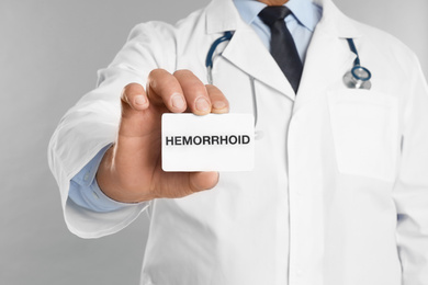 Doctor holding business card with word HEMORRHOID on light grey background, closeup