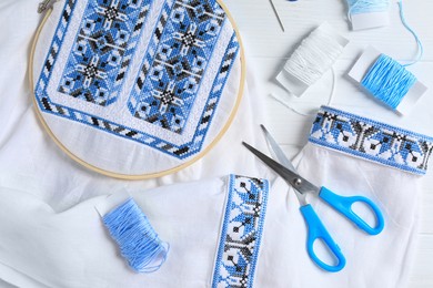 Photo of Shirt with blue embroidery design in hoop, needle, scissors and threads on white wooden table, flat lay. National Ukrainian clothes