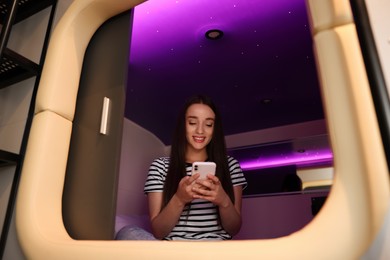 Happy young woman using smartphone in capsule of pod hostel