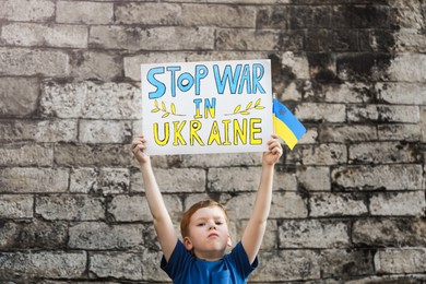 Sad boy holding poster Stop War In Ukraine and national flag against brick wall outdoors