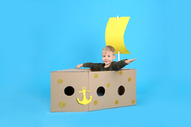 Little child playing with ship made of cardboard box on light blue background