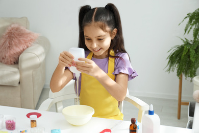 Cute little girl pouring glue into bowl at table in room. DIY slime toy