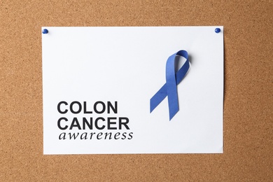 Blue ribbon and paper sheet with words COLON CANCER AWARENESS on cork background, top view
