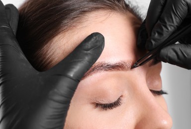Beautician tweezing woman's eyebrow on light background, closeup