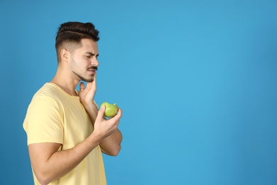Emotional young man with sensitive teeth and apple on color background. Space for text