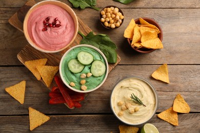 Different kinds of tasty hummus served with nachos on wooden table, flat lay