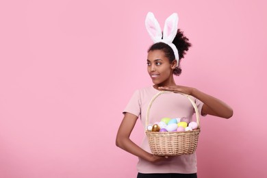 Photo of Happy African American woman in bunny ears headband holding wicker basket with Easter eggs on pink background, space for text