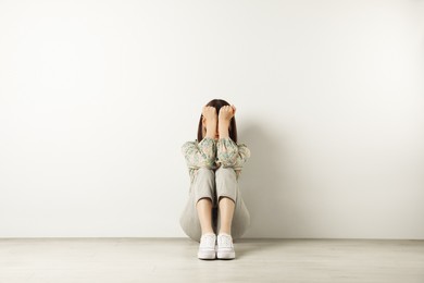 Young girl hiding face in hands on floor near white wall
