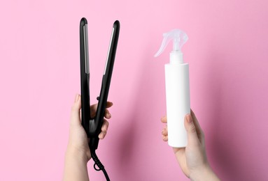 Photo of Woman holding spray bottle with thermal protection and stylish straightener on light pink background, closeup