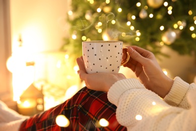 Woman with cup of hot drink near Christmas tree indoors, closeup